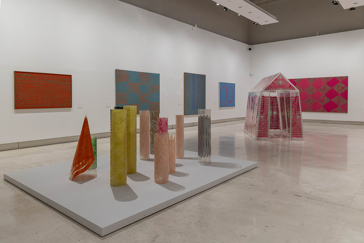 On a plinth, colorful paint lines a dozen clear tubes and cones. On the wall behind them there are 5 colorful abstract paintings covered in squiggles of contrasting colors. In the corner, a clear house-shaped plexiglass form is painted pink.