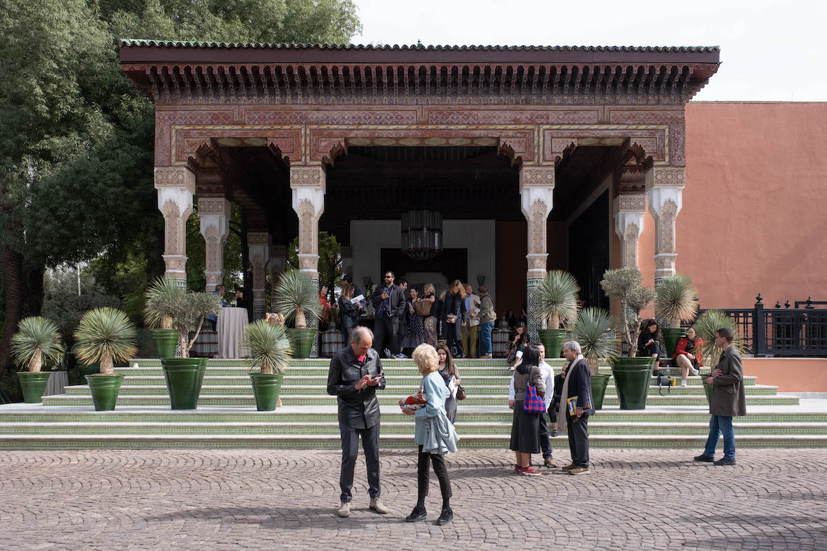 People milling around the entrance to a columned building with an ornate roof. Potted plants line the building's green stairs.