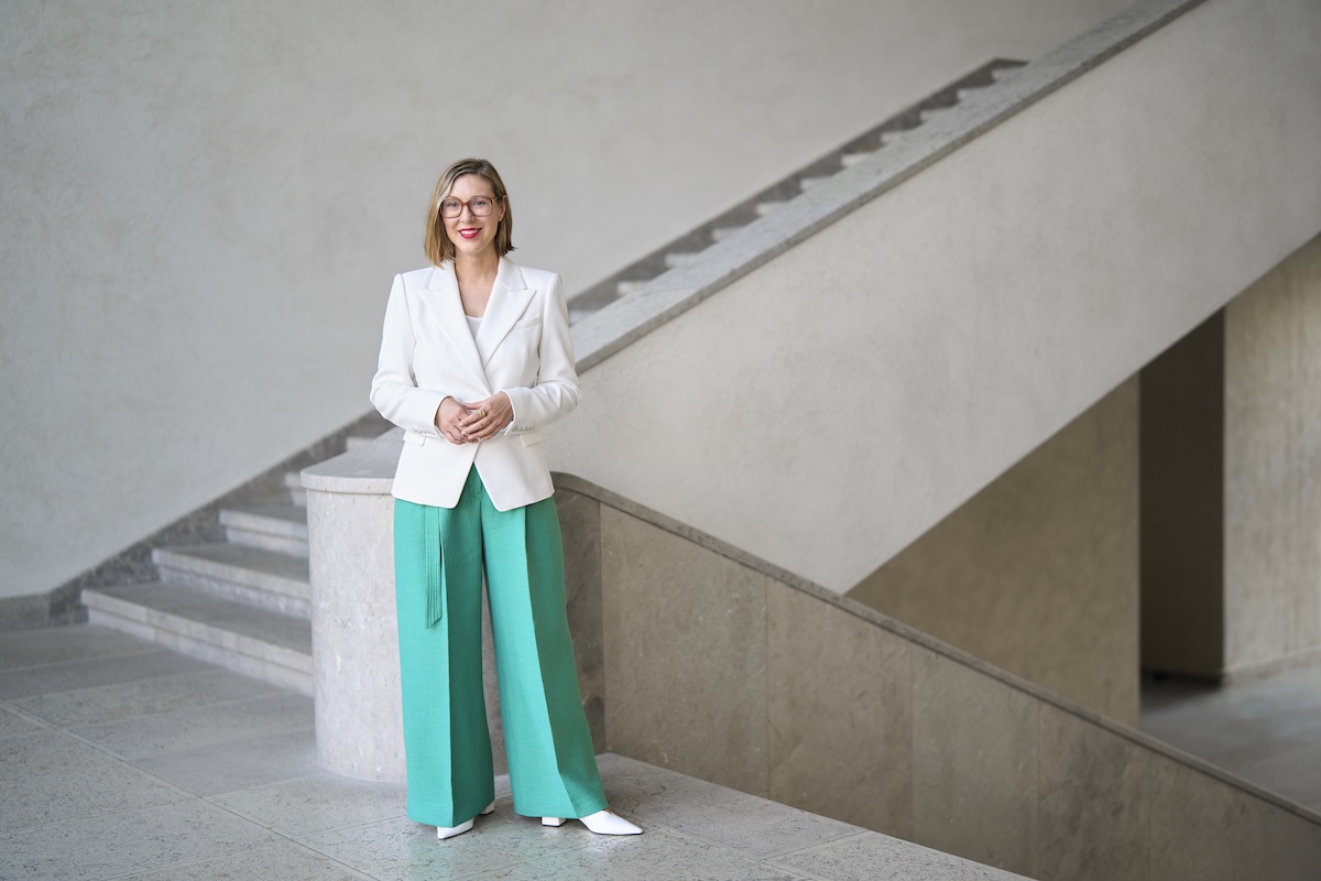 A smiling woman in a white jacket and blue pants standing beside a staircase.