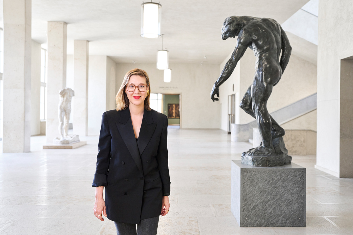 Portrait of Elena Filipovic, a white woman wearing a black double breasted blazer, as she walks through the lobby of a museum, near a Rodin sculpture.