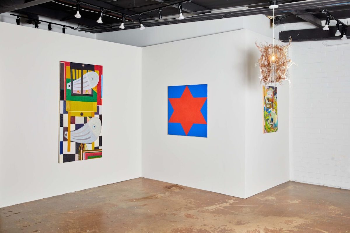 A gallery hung with a painting resembling a red star against a blue background, a painting of two figures weaving their way through a table, and more.