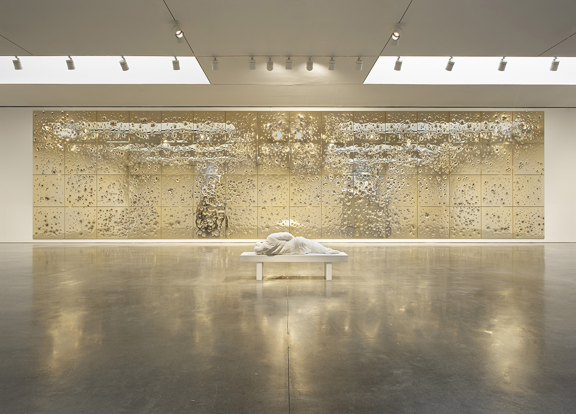 A white marble figure lies down on a bench in front of a gold plated wall covered in bullet holes.