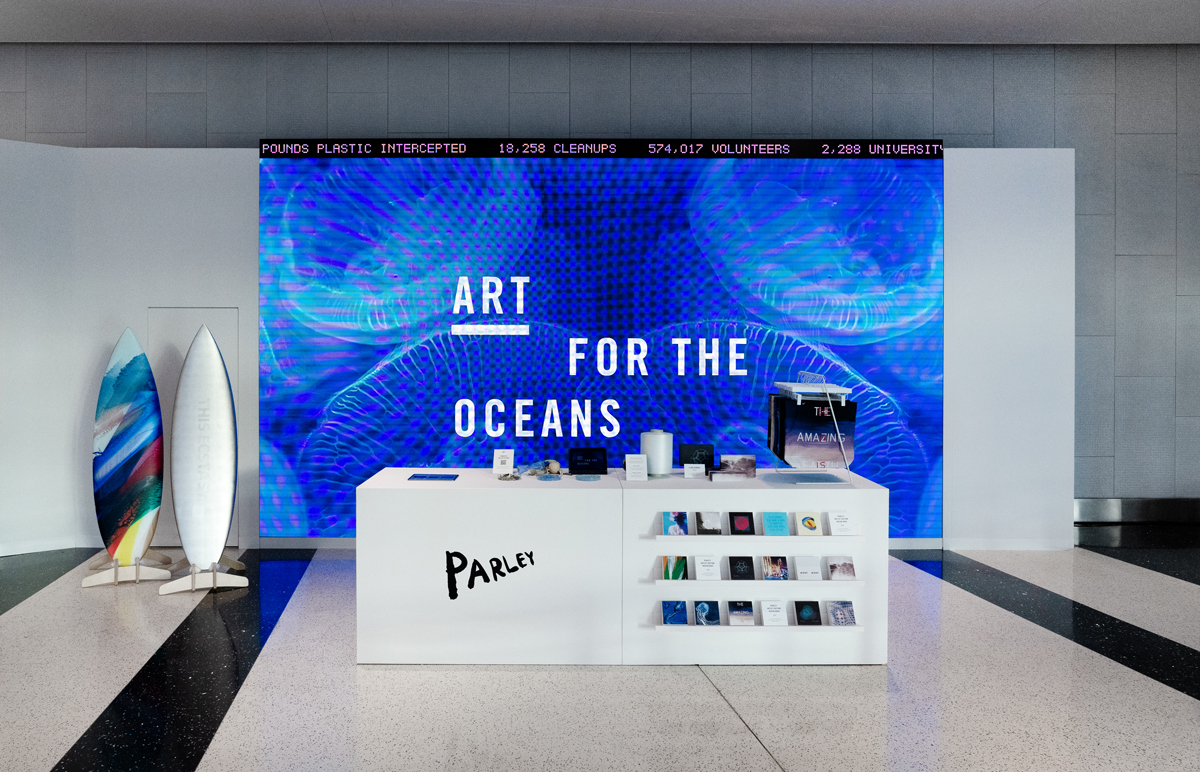 View of an LED screen that reads 'Art for the Oceans' with a table in front that reads 'Parley' and has several things on it. to the left are two artist-designed surfboards.