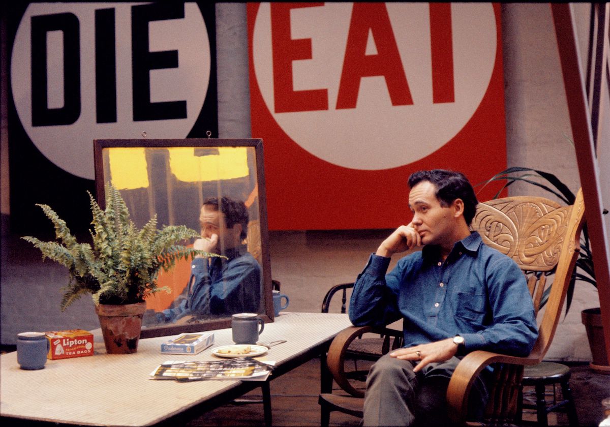 A portrait of a man seated in a wooden chair with his reflection in a mirror and two paintings on a wall with text reading "DIE" and "EAT."