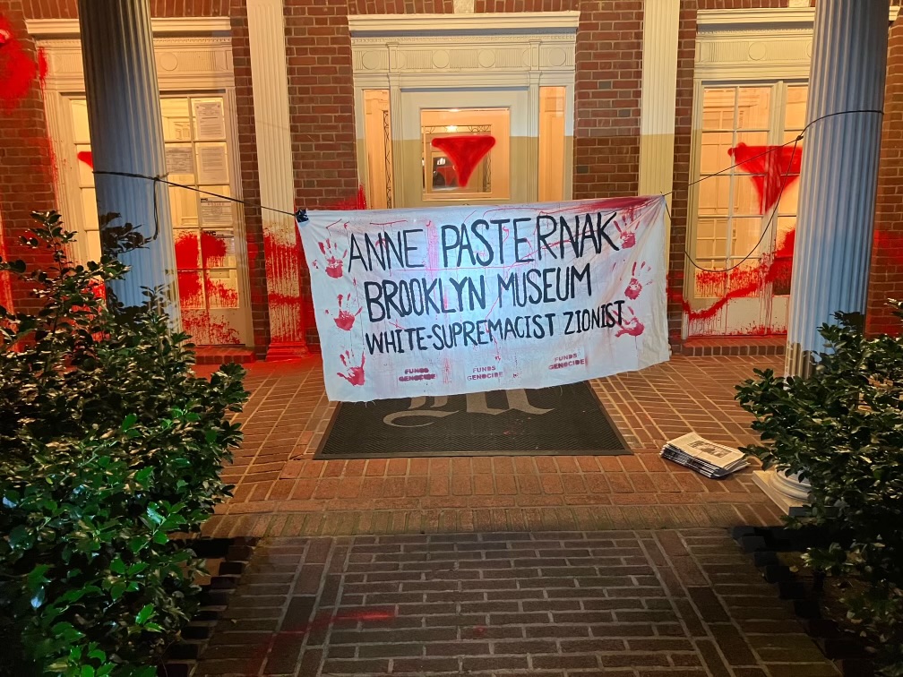 A house with a banner tied to its two columns that reads 'ANNE PASTERNAK / BROOKLYN MUSEUM / WHITE SUPREMACIST ZIONIST' with red handprints. Red spray-painted patterns also appear on the door and windows.