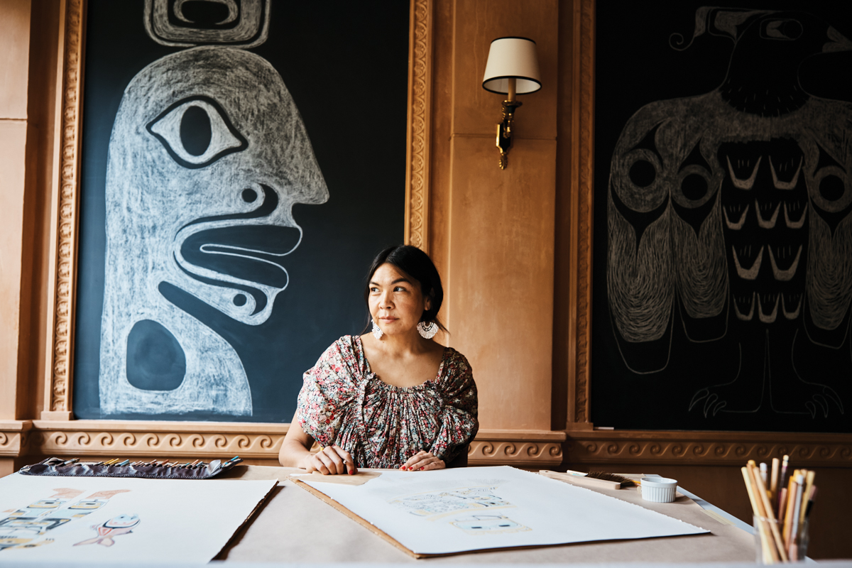 An Indigenous woman sits at t able with two drawings in front of her and two chalk murals before her.