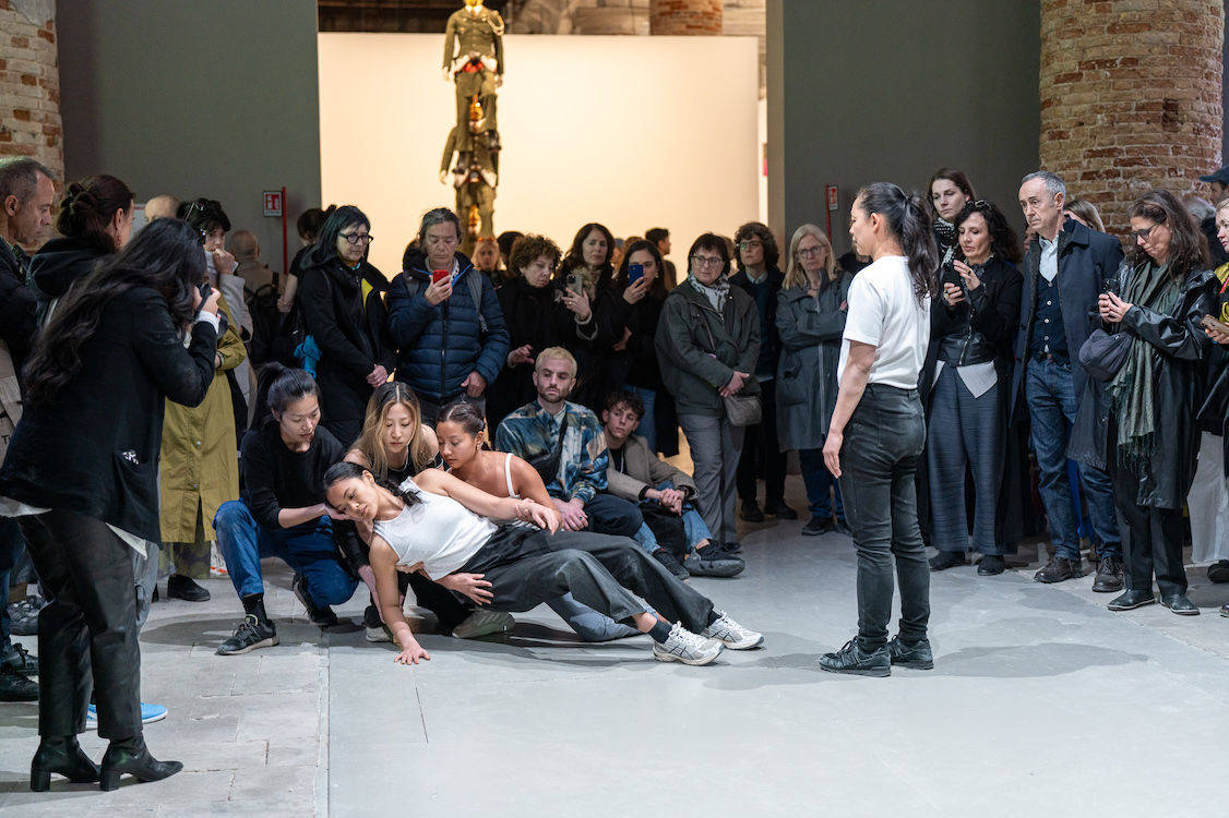 In a brick-walled gallery, a crowd watches as a falling white-shirted young woman is caught performers and gently lowered to the ground.