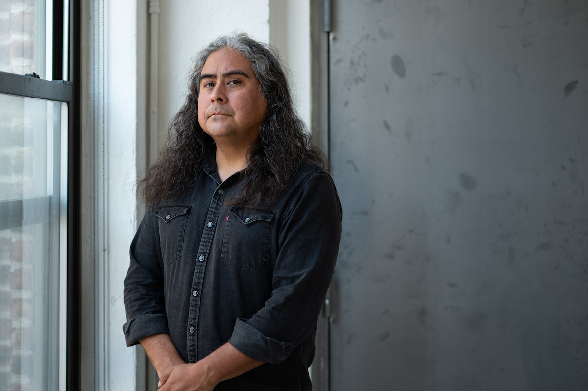 A portrait of a man with long black hair in a black denim shirt by a window.