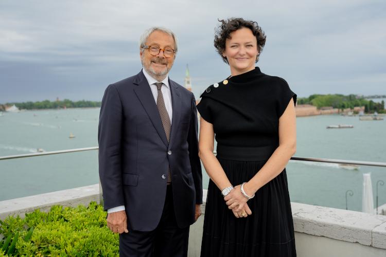 Two people—a man and a woman—stand on a rooftop overlooking a majestic harbor.