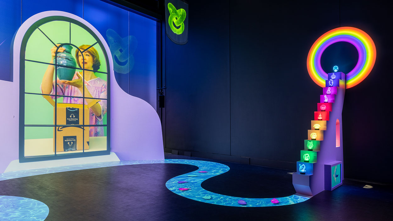 A colorful video installation shows a woman taking a vase out of an amazon box and a rainbow staircase displaying various objects.