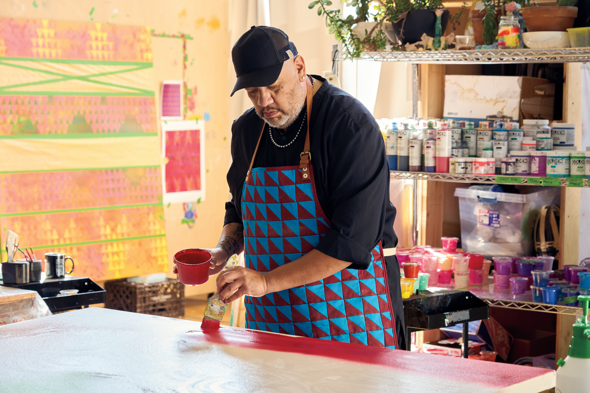 A man in a painter's smock putting a layer of paint on a canvas, in a very colorful studio.