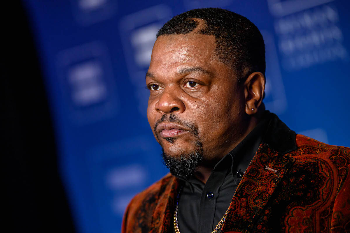 A Black man with a goatee wearing a maroon-colored jacket and a gold chain. He has a swoop cut into his hair and a stud in one ear.