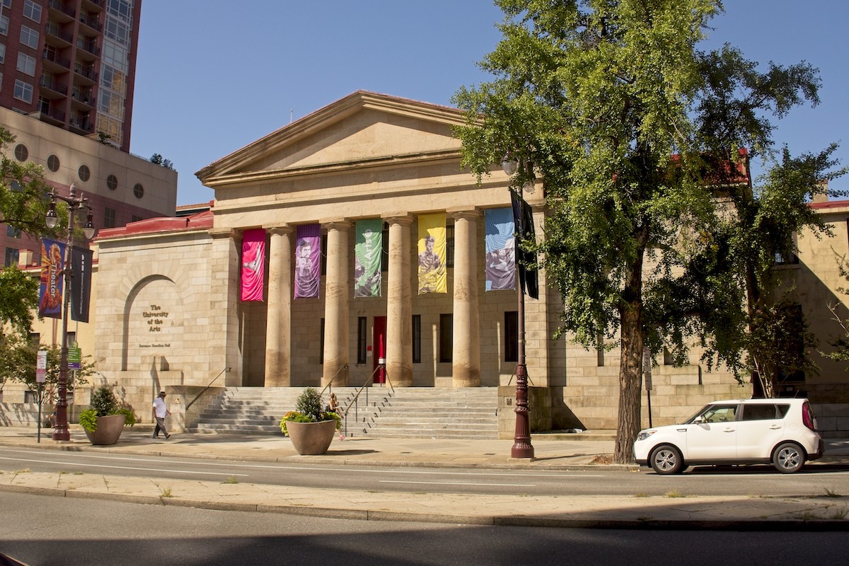 A building's columned exterior with colorful banners between each column. Beneath its stairs, a person walks by on a sidewalk.