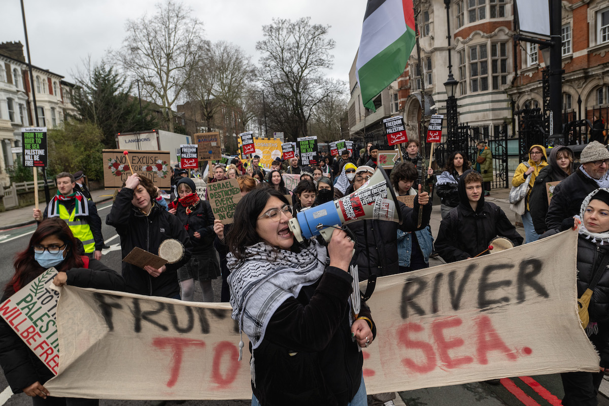 A group of people marching down a street holding a banner that reads 'FROM THE RIVER TO THE SEA.' A person yells into a megaphone near the banner. One person flies a Palestinian flag.