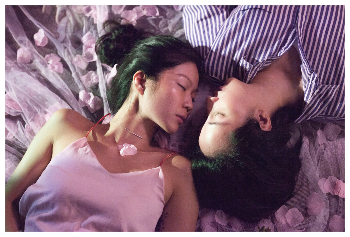 Two East Asian women are lying cheek to cheek with their eyes closed, surrounded by rose petals.