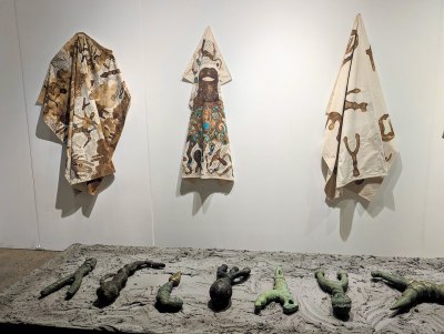 Three decorated fabrics hang on a wall. In front, a number of blunt carved objects lay on pedestal that looks covered in dried concrete or mud.