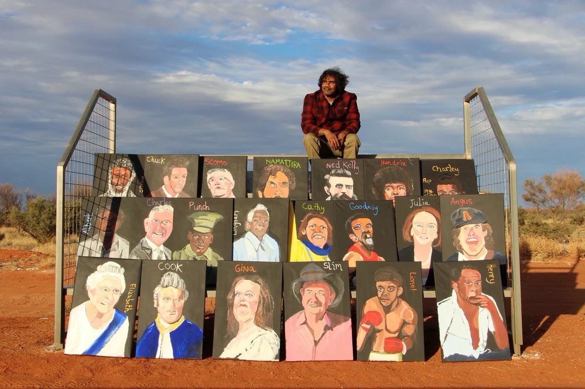 An Indigenous man seated on a riser before rows of portaits beneath him.